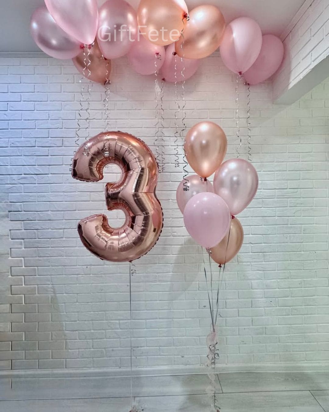 Rose gold and pink balloons with a number 3 balloon against a tiled wall.
